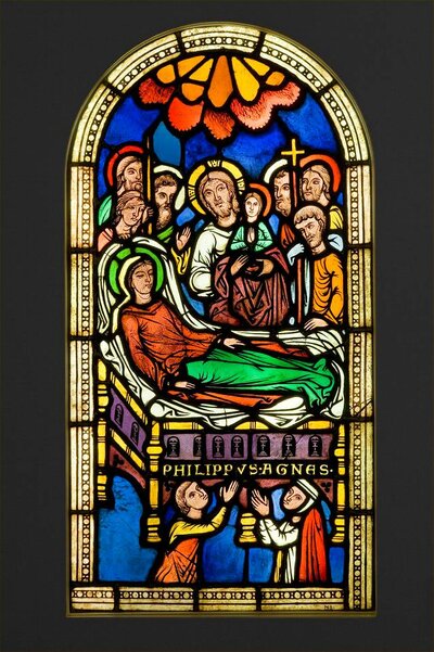 Im Zentrum der Glasmalerei liegt Maria mit geschlossenen Augen. An ihrem Bett stehen Menschen mit Jesus in ihrer Mitte. Unter ihrem Bett sind zwei betende Menschen abgebildet.