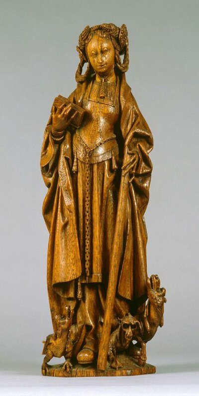 Holzfigur einer aufrechten Frau. Die rechte Hand hält ein Buch, die Linke einen Wanderstab. Zu ihren Füßen sind drei drachenähnliche Wesen.