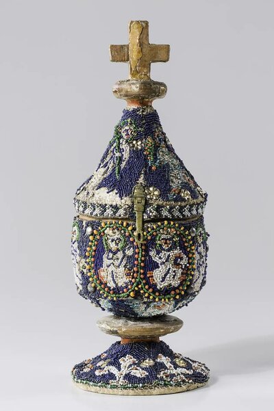 Rundliches Gefäß mit filigranen Perlenstickereien. Auf dem Deckel steht ein goldenes Kreuz.