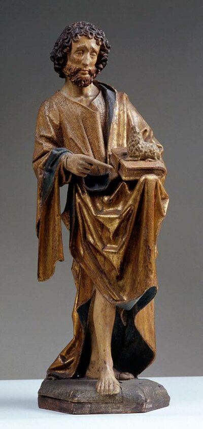 Holzfigur eines älteren Mannes. In der linken Hand hält er ein Buch, auf das er mit dem Zeigefinger der anderen Hand zeigt.