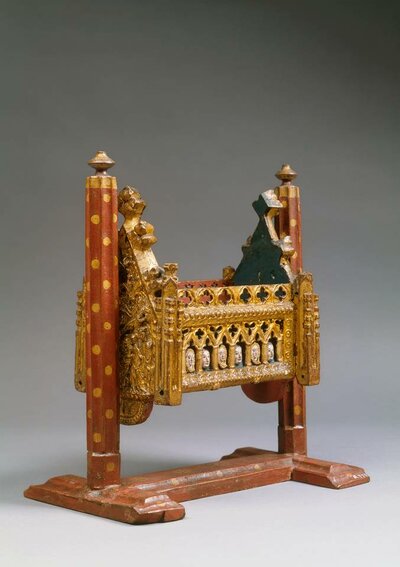 Reich verzierte rot-goldene Wiege im Stil einer gotischen Kirche mit kleinen geschnitzten Köpfen an der Seite, die durch stilisierte Spitzbogenfenster blicken