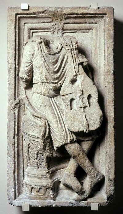 Steinrelief auf dem eine sitzende Peron mit Zupfinstrument im Arm zu sehen ist. Kopf und Hände sind abgebrochen.