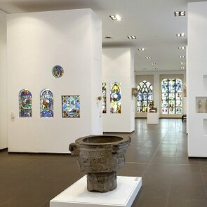 Farbfotografie eines langen Raumes mit weißen Wänden. Verschiedene Glasfenster mit Glasmalereien sind in ihm ausgestellt-