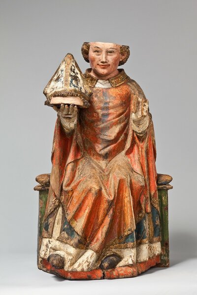 Holzfigur eines sitzenden Mannes. In seiner rechten Hand hält er seinen abgetrennten Skalp, auf dem ein Bischhofshut sitzt.