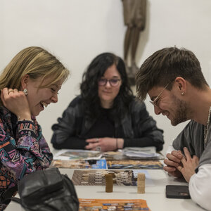 Drei Personen sitzen an einem Spieltisch. Zwischen ihnen liegt ein Spielbrett mit Figuren.