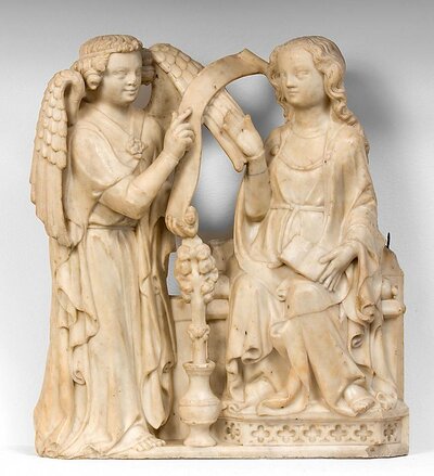 Weiße Marmorskulptur: links steht der Engel Gabriel. Er zeigt Maria, die rechts auf einem Thron sitzt, eine Stelle auf einer Schriftrolle. Zwischen beiden am Boden stehend eine Vase mit einem stilisierten Rosensproß.