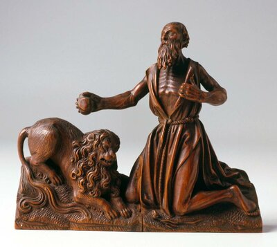 Figur aus Holz eines Mannes. Er kniet neben einem Löwen. Mit einer Hand öffnet er sein Gewand über der Brust. Sein Blick ist nach oben gerichtet.