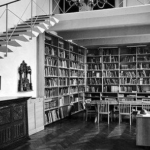 Schwarz-weiß Fotografie eines Raumes. In der Mitte steht ein großer Tisch mit Stühlen, die Wände stehen voller Regale mit Büchern.