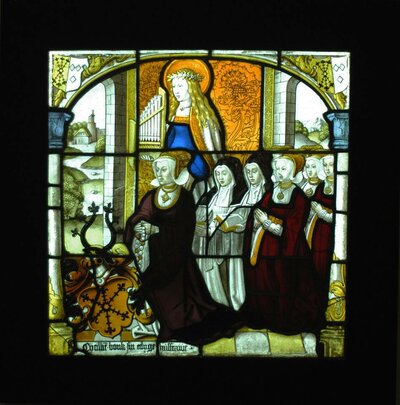 Die Glasmalerei zeigt mehrere betende Frauen. Über ihnen steht eine Frau mit Heiligenschein. In ihrer Hand hält sie eine Orgel.