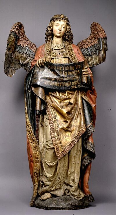 Holzfigur eines bunt bemalten weiblichen Engels. In ihrer Hand hält sie ein Spruchband.