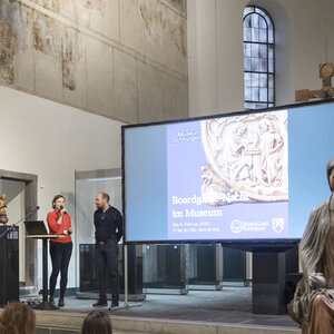 Zwei Personen stehen vor einer Beamer-Leinwand im Ostchor der Cäcilienkirche. Auf der Leinwand ist eine Folie eingeblendet mit der Aufschrift "Boardgame-Nacht im Museum".