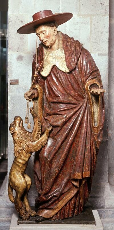 Holzfigur eines stehenden Mannes. Er trägt einen Hut mit großer Krempe und an seinem Bein springt ein kleiner Löwe hoch.