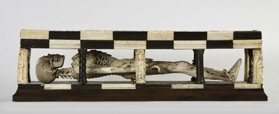 Aus Elfenbein geschnitzes Skelett eines Menschen. Dieses liegt in einer Art Sarg mit Aussparungen an den Seiten.