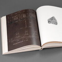 Aufgeschlagenes Buch mit einer technischen Skizze (weiß auf braunem Hintergrund, links) und einem Keil aus Stein (rechts)