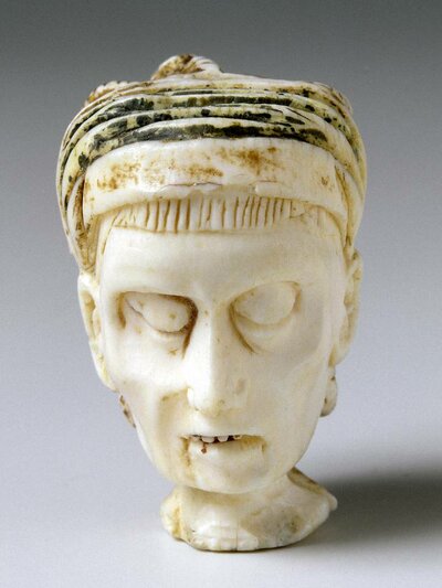 Aus Elfenbein geschnitzter Kopf. Die Augen sind geschlossen. Der Mund steht ein Stück offen sodass Zähne zu sehen sind..