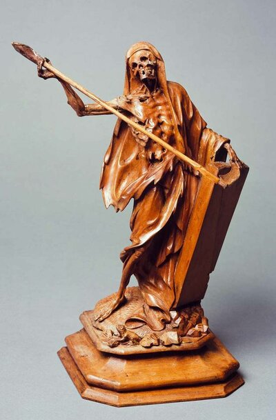 Statue aus Holz mit einem stehenden, in Lumpen gehüllten Skelett, einer Sense und einem Grabstein. Am Fuß sitzt eine Kröte.