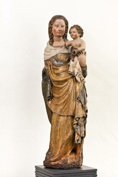 Holzfigur einer stehenden Maria. Auf ihrem Arm hält sie das Jesuskind. Ihr Gewand ist braun bemalt.