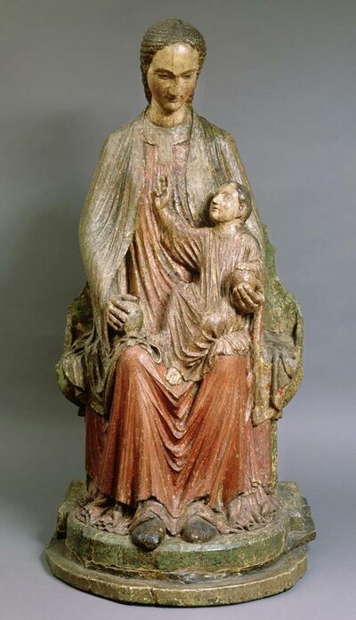 Holzfigur einer sitzenden Frau mit gesenktem Kopf. Auf ihrem Schoß sitzt ein Kind, das zu ihr aufsieht und die rechte Hand gehoben hat.