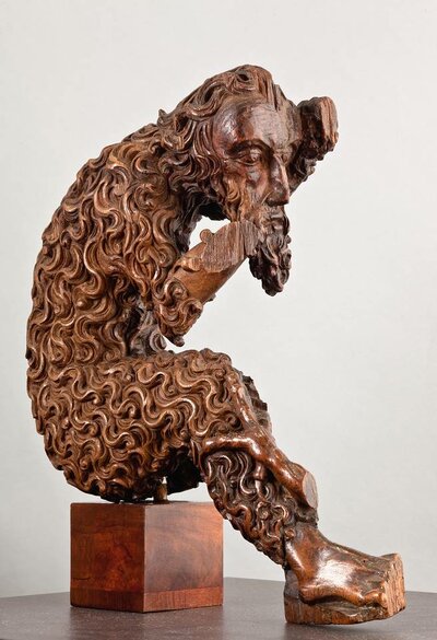 Ein aus Holz geschnitzter Mann mit Bart und kantigem Gesicht sitzt auf einem Holzwürfel. In seinen hölzernen Körper sind überall Locken eingraviert. 