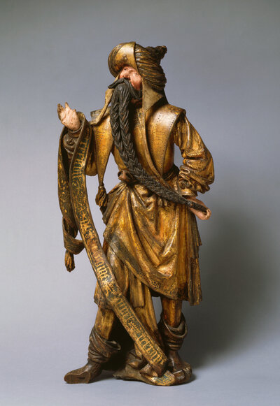 Holzfigur eines stehenden Mannes mit Turban und sehr langem geflochtenen Bart. Sein Blick ist nach oben gerichtet. Über seinem gehobenen Arm liegt ein Schal.