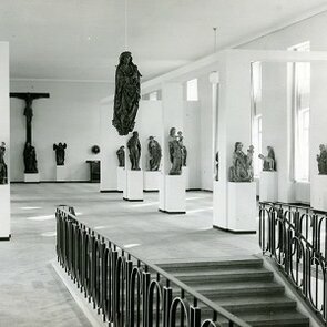 Schwarz-weiß Fotografie von einem langen Raum mit hohen Decken, in dem verschiedene Skulpturen ausgestellt sind.