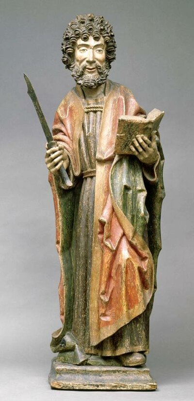 Bunte Holzfigur eines stehenden Mannes: In der rechten Hand hält er ein kurzes Schwert. Die linke Hand hält ein aufgeschlagenes Buch.