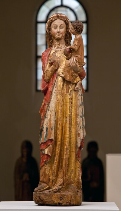 Stehende Madonna in einem goldenen Gewand und einem roten Mantel mit dem sitzenden Jesukind auf dem linken Arm. Das Kind blickt auf das Gesicht der Madonnenfigur, die Madonna blickt auf den Betrachter. Die rechte Hand der Madonna fehlt.