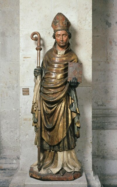 Holzfigur eines stehenden Mannes mit Bischofsstab und Bischofshut. Seine linke Hand hält ein Buch.