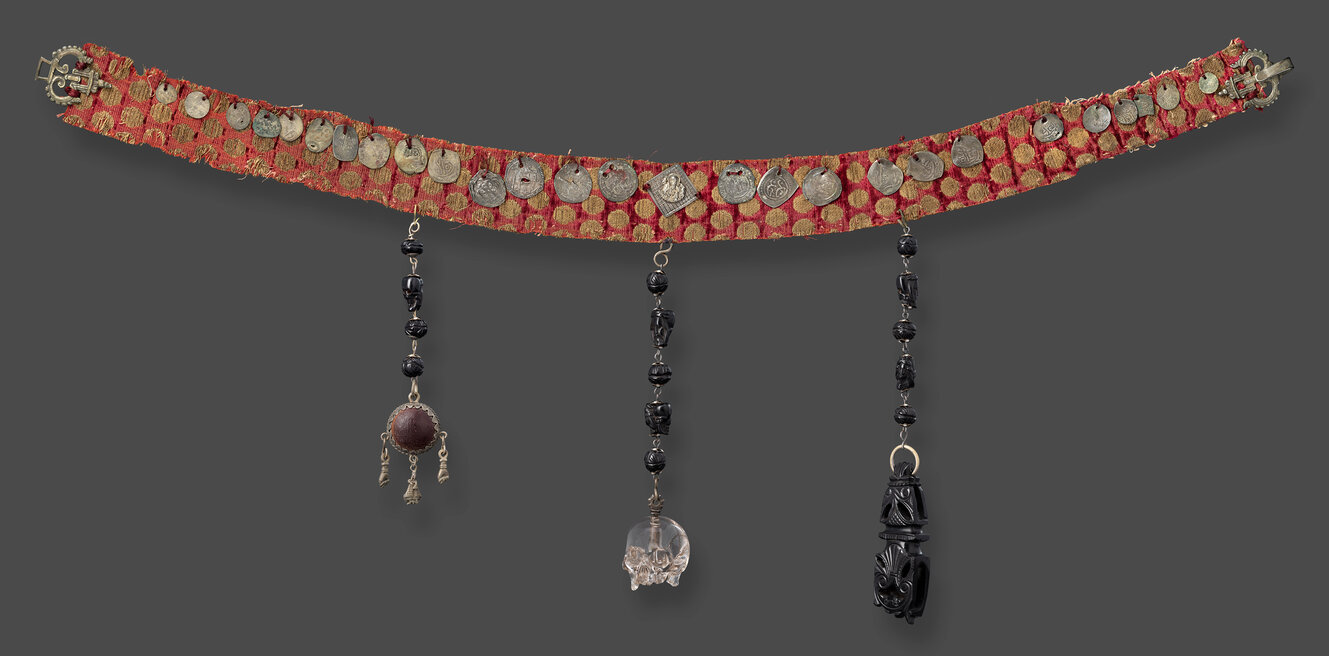 Gürtel aus rotem Stoff mit Goldfäden durchwirkt und mit Münzen besetzt. Daran hängen drei Gliederketten mit geschitzten schwarzen Perlen und einem Totenschädel aus Bergkristall.