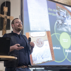 Ein Mann steht vor einer Beamer-Leinwand und hält eine Präsentation. In seiner Hand hält er ein Mikrofon.