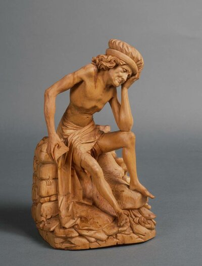 Holzfigur eines sitzenden Mannes. Seinen Kopf hat er in seine linke Hand gelegt. Sein Blick ist zum Boden gesenkt.