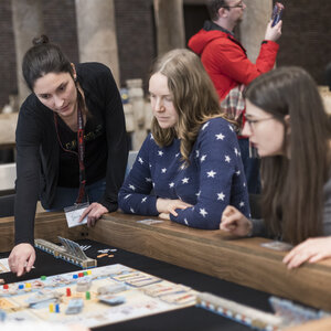 Eine Grupppe Frauen sitzt an einem Brettspieltisch. Eine Frau erklärt die Spielregeln.