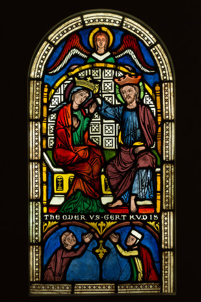 Glasmalerei, die abbildet wie Jesus Maria eine Krone aufsetzt. Über ihnen ist ein Engel abgebildet, unter ihnen zwei betende Menschen.