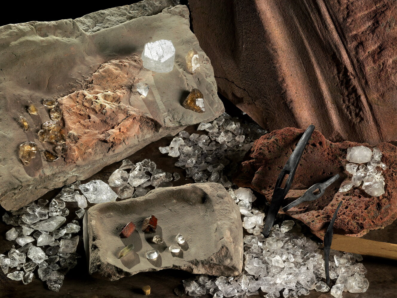 Verschiedene geschliffene und ungeschliffene Bergkristalle und Werkzeug zur Bearbeitung liegen auf verschiedenen Untergründen aus Stein, Ton und Brot