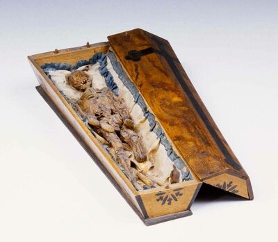 Wachstödlein auf Seidenstoff in einem Holzsarg.